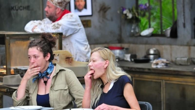 Leila Lowfire und Sibylle Rauch essen Dschungelpizza vor einem sichtlich gelangweilten Koch. (Bild: RTL)