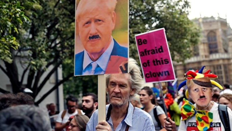 Diese Demonstranten sehen Premier Boris Johnson als Clown mit Hitler-Schnurrbart. (Bild: AP)