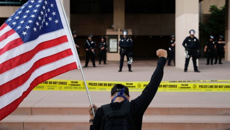 Ein Demonstrant kniet mit der US-Flagge in der Hand vor der Polizei, die das Rathaus der kalifornischen Stadt Anaheim bewacht. (Bild: AP)