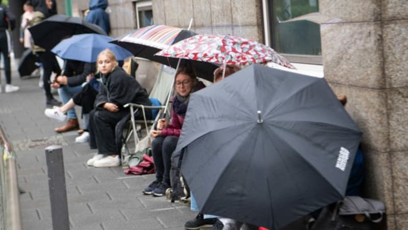 Journalisten und Prozessbesucher warten vor dem Gerichtsgebäude mit Regenschirmen und Campingsesseln auf Einlass. (Bild: APA/dpa/Boris Roessler)