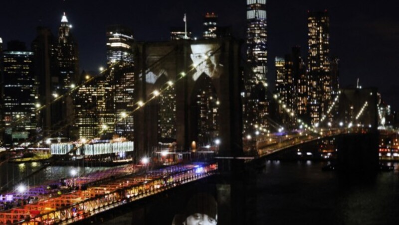 (Bild: NEW YORK, NEW YORK - MARCH 14: Faces of victims of Covid-19 are projected onto the Brooklyn Bridge during a memorial service called A COVID-19 Day of Remembrance on March 14, 2021 in New York City. The event, which will includes a virtual performance by The New York Philharmonic, marks the day the first known coronavirus fatality was confirmed in the city. Spencer Platt/Getty Images/AFP (Photo by SPENCER PLATT / GETTY IMAGES NORTH AMERICA / Getty Images via AFP))