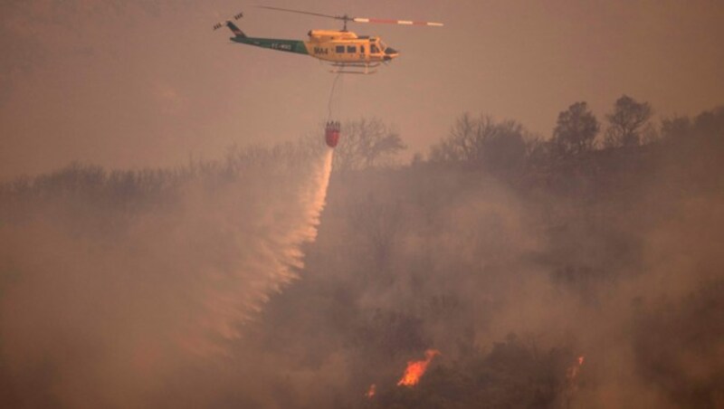 Ein Hubschrauber wird von Alhaurin de la Torre aus fotografiert, als er Wasser über einem Waldbrand in der Sierra de Mijas in der Provinz Malaga abwirft. (Bild: APA/Photo by JORGE GUERRERO/AFP)