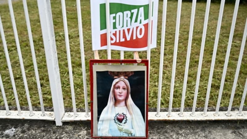 Anhänger des ehemaligen Ministerpräsidenten haben ein Bild von der Jungfrau Maria und ein Schild mit der Aufschrift: „Vorwärts, Berlusconi“ am Zaun des Spitals San Raffaele aufgestellt. (Bild: APA/AFP/GABRIEL BOUYS)