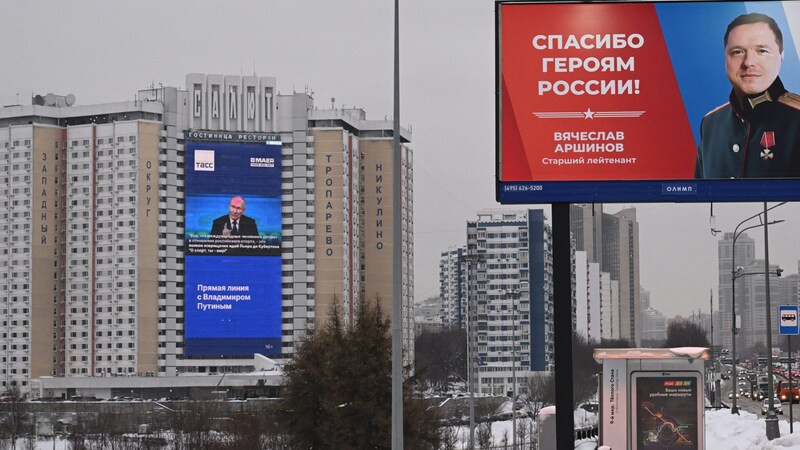 Die Jahreskonferenz des russischen Präsidenten Wladimir Putin wird an einer Häuserfassade übertragen. Rechts davon wird ein russischer Soldat auf einem Plakat geehrt. (Bild: APA/AFP/NATALIA KOLESNIKOVA)