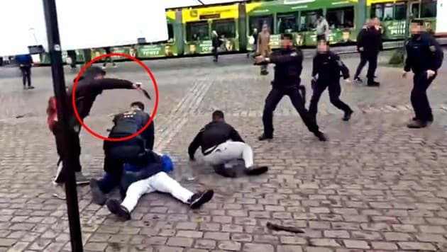 Sulaiman A. setzt mit dem Kampfmesser zum ersten Stich in den Hals von Rouven L. an, der gerade den Mann in der blauen Jacke festhält. Der hatte irrtümlich auf einen Helfer (rechts) eingeschlagen, der den Attentäter fixiert hatte. (Bild: Quelle: YouTube/Augen auf!; krone.at)