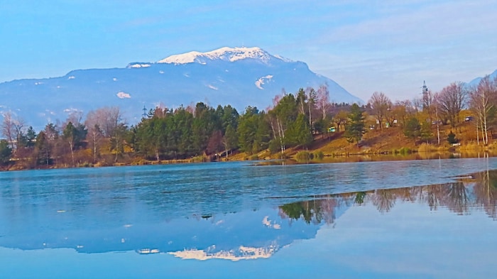 Blue sky and clouds are reflected in the only partially frozen Silbersee lake near Villach, by ...
