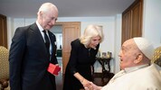 Charles und Camilla sind auf dem Foto schwarz gekleidet und lächeln. Der König hält ein rotes ...