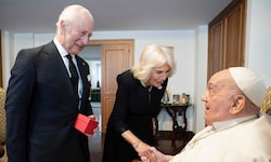 Charles und Camilla sind auf dem Foto schwarz gekleidet und lächeln. Der König hält ein rotes ...