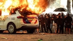 Ein wütender und zum Teil vermummter Mob sieht zu, wie ein Polizeifahrzeug abbrennt.