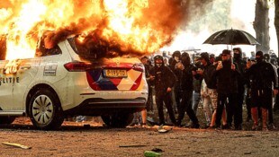 Ein wütender und zum Teil vermummter Mob sieht zu, wie ein Polizeifahrzeug abbrennt.