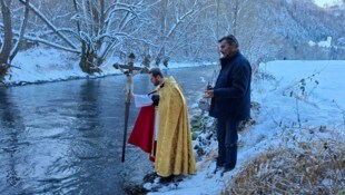 Pfarrer Bernd Wegscheider taucht das Prozessionskreuz in die Glan, Mesner Johann Karner hält ...