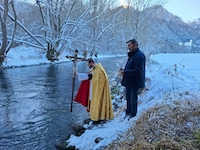 Pfarrer Bernd Wegscheider taucht das Prozessionskreuz in die Glan, Mesner Johann Karner hält ...