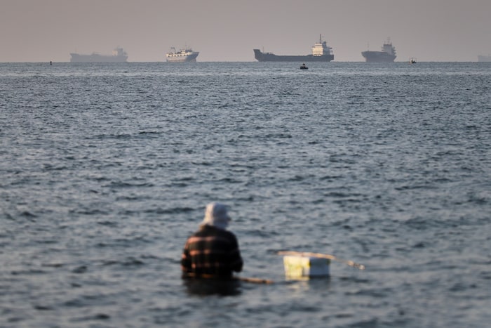 Eine Person sitzt im seichten Wasser, während Fracht- und Handelsschiffe in der Straße von ...