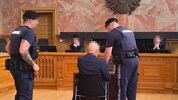 Der Angeklagte vor dem Richtersenat, bewacht von Justizwachebeamten.