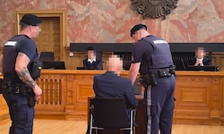 Der Angeklagte vor dem Richtersenat, bewacht von Justizwachebeamten.